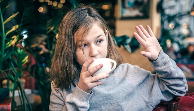 Een klein meisje met een kopje thee in een café die hallo zwaait naar iemand