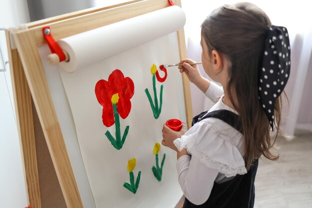 Een klein meisje met een palet in haar handen tekent bloemen op een ezel