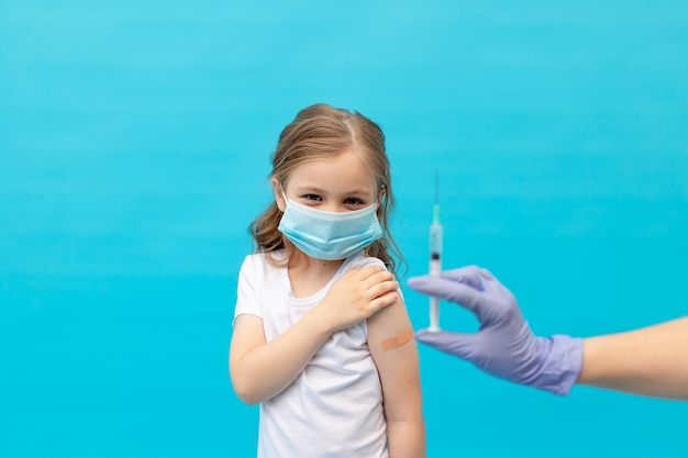 Een klein meisje met een pleister op haar arm na vaccinatie in een medisch beschermend masker op een blauwe achtergrond en de hand van een verpleegster met een spuit, ruimte voor tekst, het concept van vaccinatie