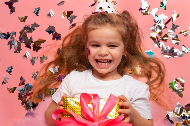 Een klein meisje met een verjaardagshoed houdt een cadeau voor een roze achtergrond met confetti.