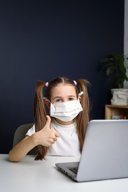 Een klein meisje met masker om covid coronavirus te beschermen en duimen te laten zien.