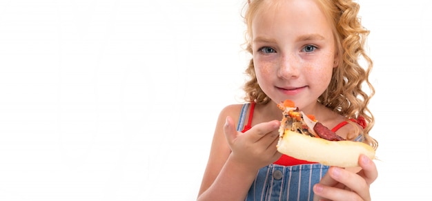 Een klein meisje met rood haar in een rode trui en een blauw-witte jumpsuit in een streep eet een groot pizzapunt