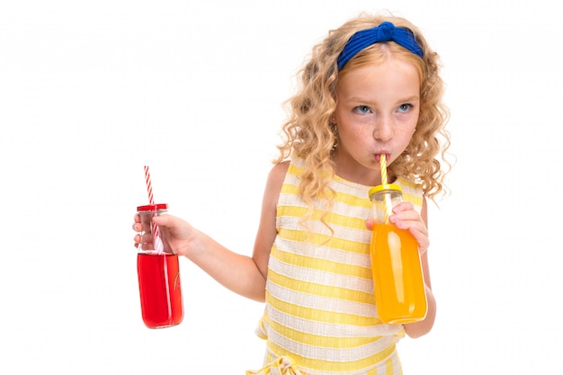 Een klein meisje met rood haar in een wit en geel gestreept zomerpak, met een blauw verband op haar hoofd, drinkt sinaasappelsap in een glazen fles pijp en houdt watermeloensap.