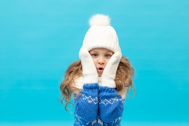 Een klein meisje raakt verrassend haar gezicht aan met haar handen op een blauwe achtergrond in een wintermuts en een trui met wanten, ruimte voor tekst