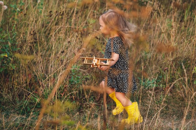 Een klein meisje speelt met een houten vliegtuig in de natuur Vrijheid droom verbeelding concept Gelukkige jeugd Een mooi meisje loopt vreugdevol langs een pad in het gras lacht glimlacht