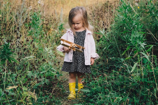 Een klein meisje speelt met een houten vliegtuig in een dorp in een veld Kinder geluk gelukkige jeugd concept Een mooi blond meisje in een jurk en laarzen ziet er bedachtzaam uit