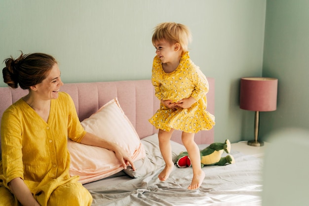 Een klein meisje springt vrolijk op het bed, moeder lacht en gaat naast haar zitten.