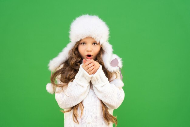 Een klein meisje verwarmt haar handen door warme lucht van achter haar mond te blazen. een model met krullend haar en een warme trui. Een kind in warme kleren in het winterseizoen op een geïsoleerde achtergrond.