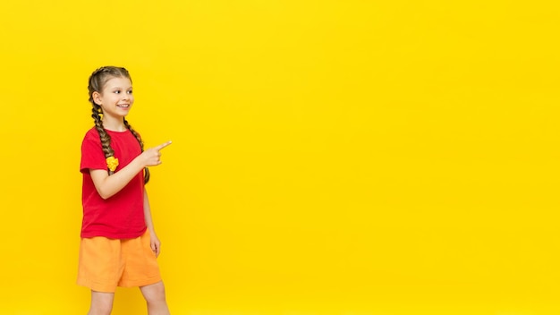 Een klein meisje wijst met haar vinger naar een lege ruimte voor uw advertentie op een gele geïsoleerde achtergrond Een mooi kind lacht in een rood T-shirt met twee staartjes Kopieer ruimte