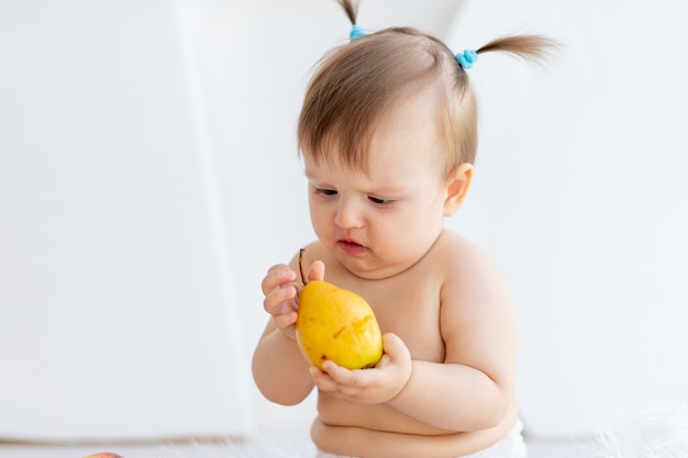 Een klein meisje zit in een lichte kamer met een bord fruit en eet een peer