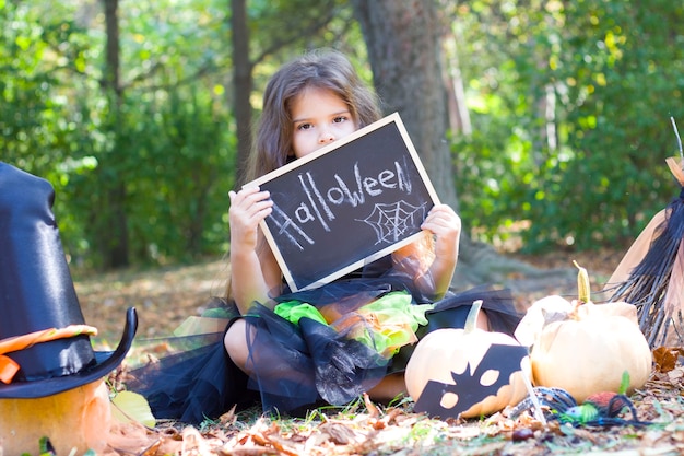 Een klein meisje zit in een park met gele bladeren in een heksenkostuum. Bezem, pompoen, vleermuismasker, heksenhoed. Halloween.