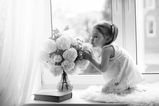 Een klein meisje zit op de vensterbank. Een boeket bloemen in een vaas bij het raam en een meisje dat bloemen snuffelt. Een prinsesje in een witte jurk met een boeket witte bloemen bij het raam.