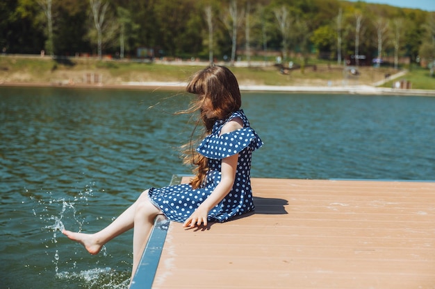Een klein meisje zit op een pier in de buurt van het stadsmeer in de buitenlucht zomervakantie