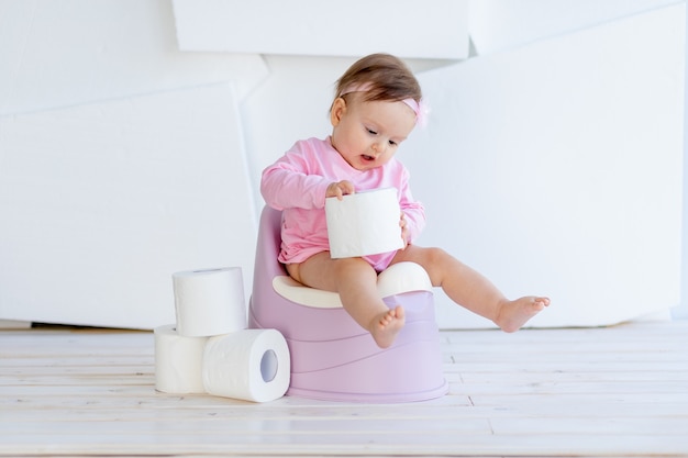 Een klein meisje zit op een potje in roze kleren in een lichte kamer en speelt met wc-papier