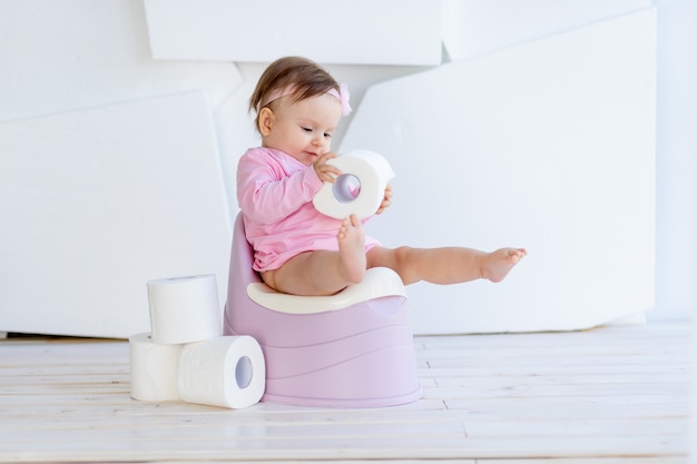 Een klein meisje zit op een potje in roze kleren in een lichte kamer en speelt met wc-papier