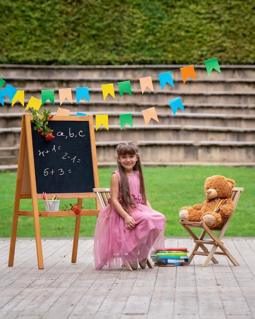 Een klein meisje zit op een stoel naast een teddybeer tegen de achtergrond van een schoolbestuur