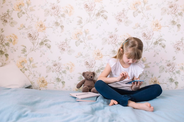 Foto een klein meisje zit op het bed in de stijlvolle slaapkamer met een telefoon in haar hand en leest in een smartphone