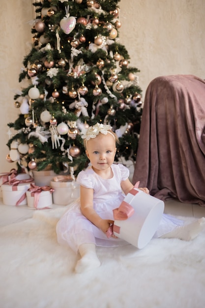 Een klein mooi meisje in een feestelijke witte jurk zit op een deken in de buurt van de kerstboom met een cadeau