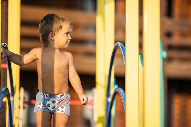 Een klein naakt vrolijk kind in felblauwe korte broek speelt op een kinderspeelplaats terwijl hij buiten loopt op een warme zonnige dag onder de gouden zomeravondzon