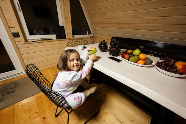 Een klein schattig babymeisje in een pyjama zit in de keuken met groene appel in een gezellig houten huisje in een kleine hut. Leven op het platteland