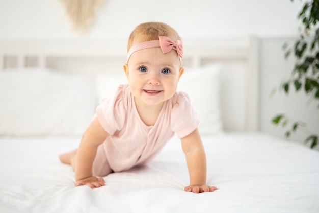 Een klein schattig gezond meisje tot een jaar oud in een roze bodysuit gemaakt van natuurlijke stof zit op een bed op wit beddengoed in de slaapkamer kijkend naar de camera de baby is thuis