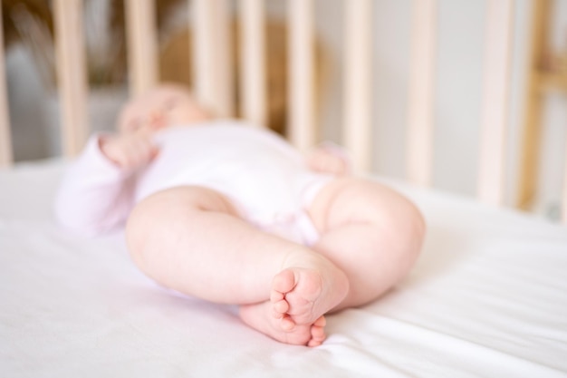 Een klein schattig glimlachend babymeisje in een bodysuit ligt op het bed in de slaapkamer van het huis op wit beddengoed op haar achterpoten van een gezonde babyclose-up