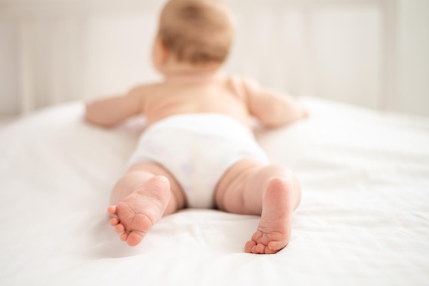 Een klein schattig glimlachend babymeisje naakt in wit slipje ligt op het bed in de slaapkamer van het huis op wit beddengoed op haar buik benen van een gezonde baby close-up