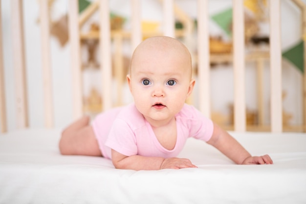 Een klein schattig glimlachend meisje in een roze bodysuit ligt in een wieg thuis op wit beddengoed op haar buik kijkend naar de camera glimlachende gelukkige baby