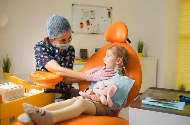 Een klein schattig glimlachend meisje zit in een oranje tandartsstoel en toont een vrouwelijke tandarts een zere tand