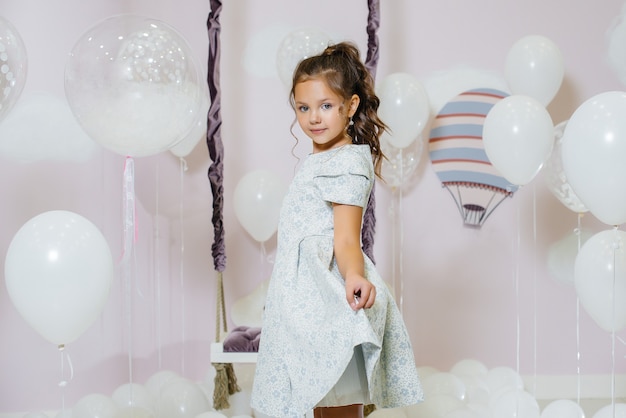Een klein schattig meisje glimlacht en poseert tegen de muur van een schommel met ballonnen.