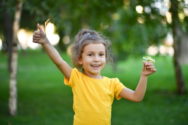 Een klein schattig meisje in een geel T-shirt tegen de achtergrond van groene bomen