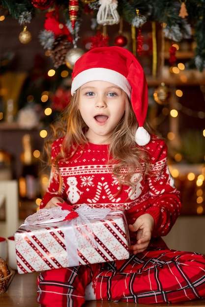 Een klein schattig meisje in een rode trui en een kerstmuts in een donkere keuken met een kerstboom verheugt zich met geschenken en wacht op het nieuwe jaar of kerstmis