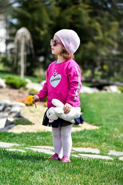 Een klein schattig meisje in een roze jasje en roze hoed loopt door de stad. Het meisje houdt een paardenbloem in de ene hand, een speeltje in de andere hand. Het is zonnig buiten. Lente. Zonnig. Gelukkige jeugd.