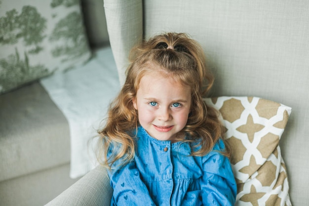 Een klein schattig meisje met blauwe ogen en krullend lang haar zit thuis in een gezellige fauteuil Gezellig licht huis Gelukkig kind rust