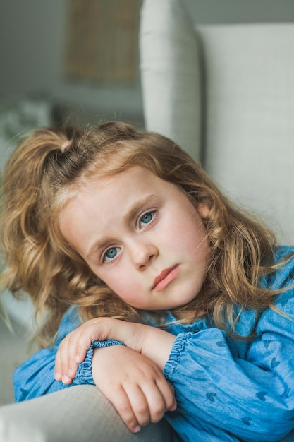 Een klein schattig meisje met blauwe ogen en krullend lang haar zit thuis in een gezellige fauteuil Gezellig licht huis Gelukkig kind rust