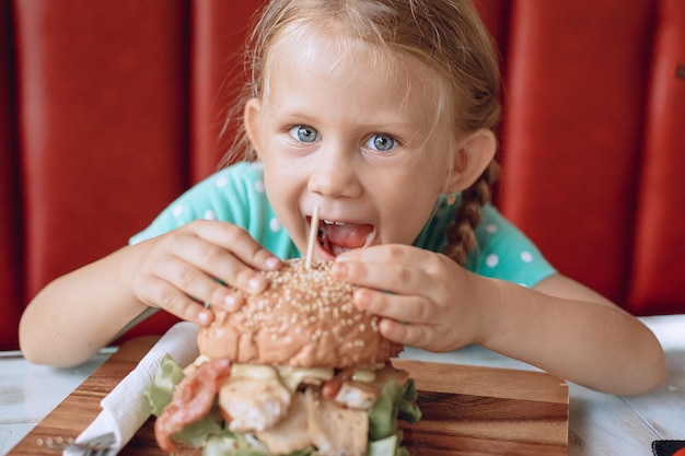 Een klein schattig meisje met blond haar en blauwe ogen probeert een grote burger te bijten in een plaatselijk café. Portret.