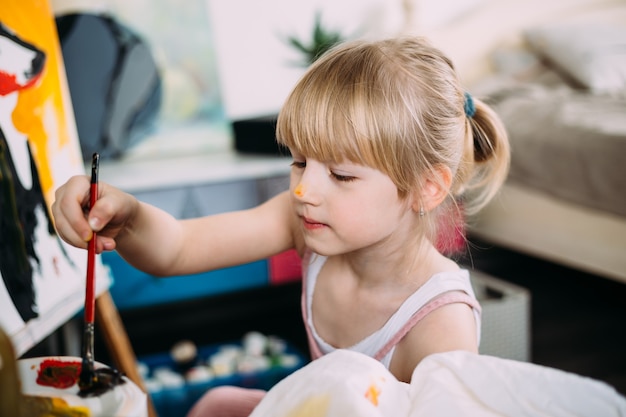 Een klein schattig meisje schildert thuis een grote foto met acryl op een ezel