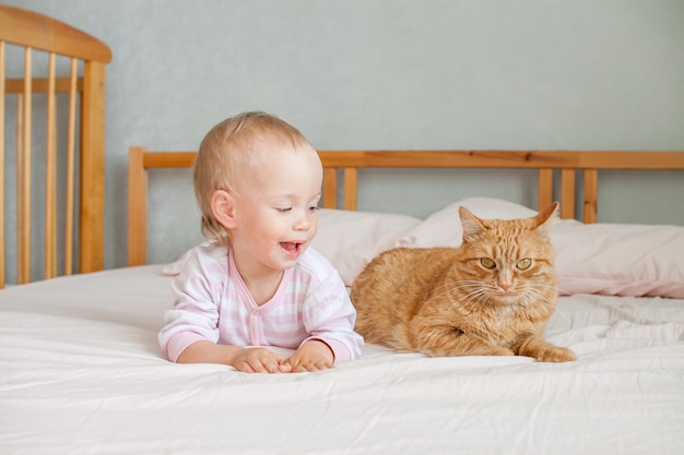 Een klein schattig meisje zit op het bed met een dikke gemberkat streelt en speelt met hem