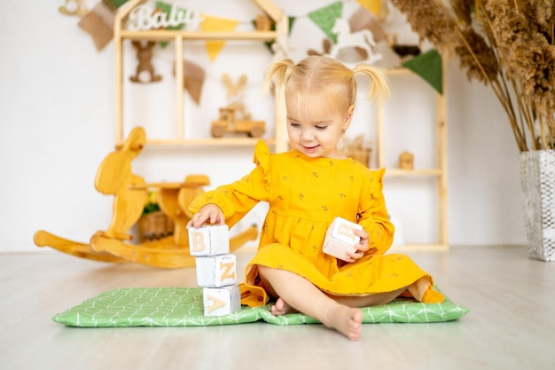 Een klein schommelend babymeisje staat een piramide of een toren van houten kubussen in een speelhuisje en lacht een gelukkig kind in een gele jurk