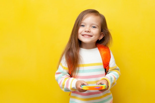 Een klein schoolmeisje in een gestreepte blouse en met een rugzak houdt een pop in haar handen en glimlacht