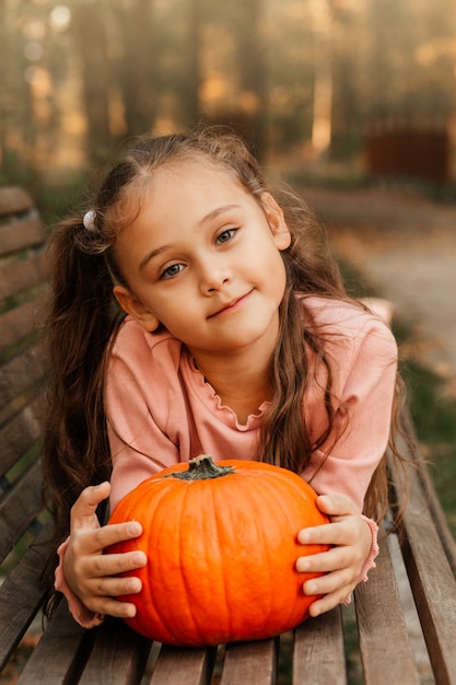 Een klein vrolijk meisje houdt een oranje pompoen in het park in de herfst. Halloween-rekwisieten