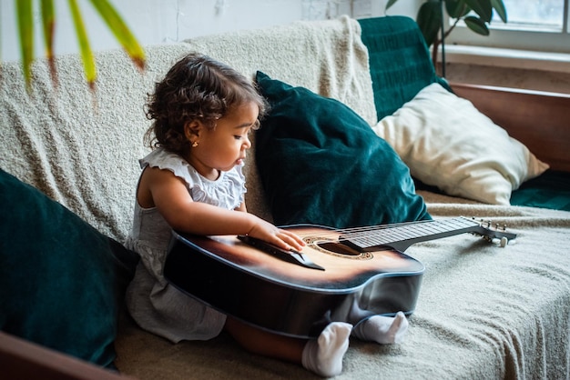 Een klein zwart meisje met een hoed zit op een bank en speelt gitaar, baby speelt, componeert melodie en muziek