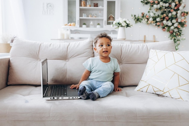 Een kleine Afro-Amerikaanse jongen met een donkere huidskleur zit op een bank met een laptop