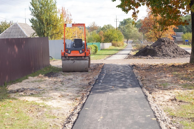 Een kleine bestratingsafwerkmachine staat in de herfst in de buurt van een nieuw asfaltpad Wegreparaties