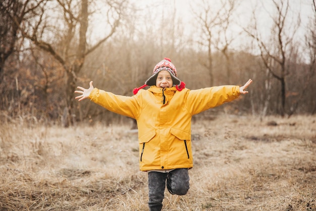 Een kleine gelukkige jongen in een gebreide muts en een geel jasje loopt op een zonnige dag langs een pad in het herfstpark.