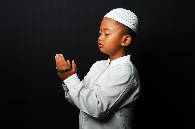 Een kleine jongen die een pet draagt, bidt geïsoleerd op zwarte achtergrond