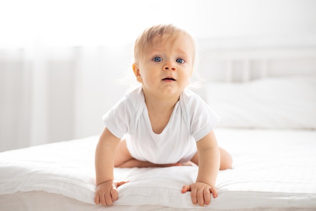 Foto een kleine jongen een blond kind met blauwe ogen in een wit bodysuit is op alle vier thuis in de slaapkamer op een bed met witte laken en kijkt naar de camera