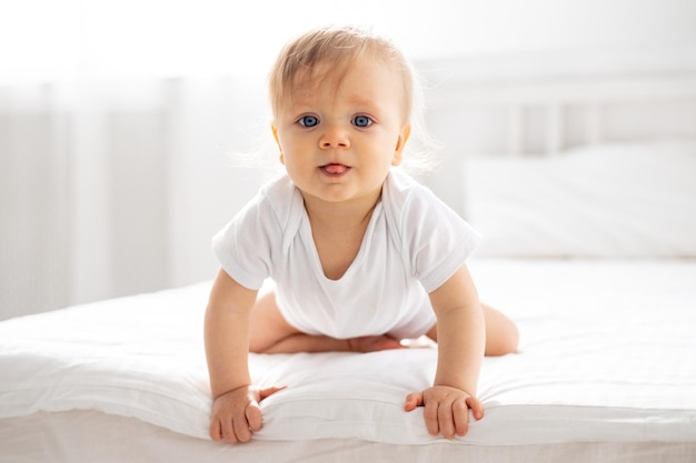 Foto een kleine jongen, een blondharig kind met blauwe ogen in een witte bodysuit, zit op handen en voeten thuis in de slaapkamer op een bed met wit beddengoed en kijkt naar de camera