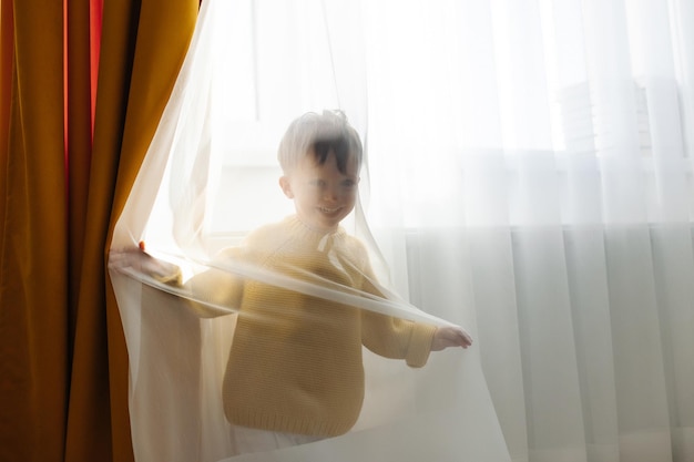 Een kleine jongen in een geel jasje verstopt zich voor zijn moeder achter een gordijn in de kinderkamer