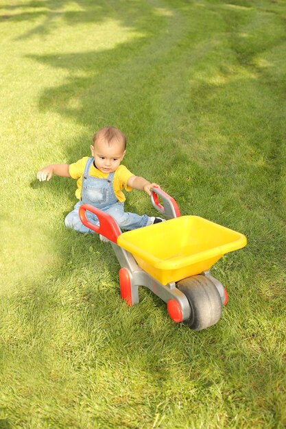 een kleine jongen in een geel T-shirt en spijkerbroek speelt op het grasveld in het park met een gele kar